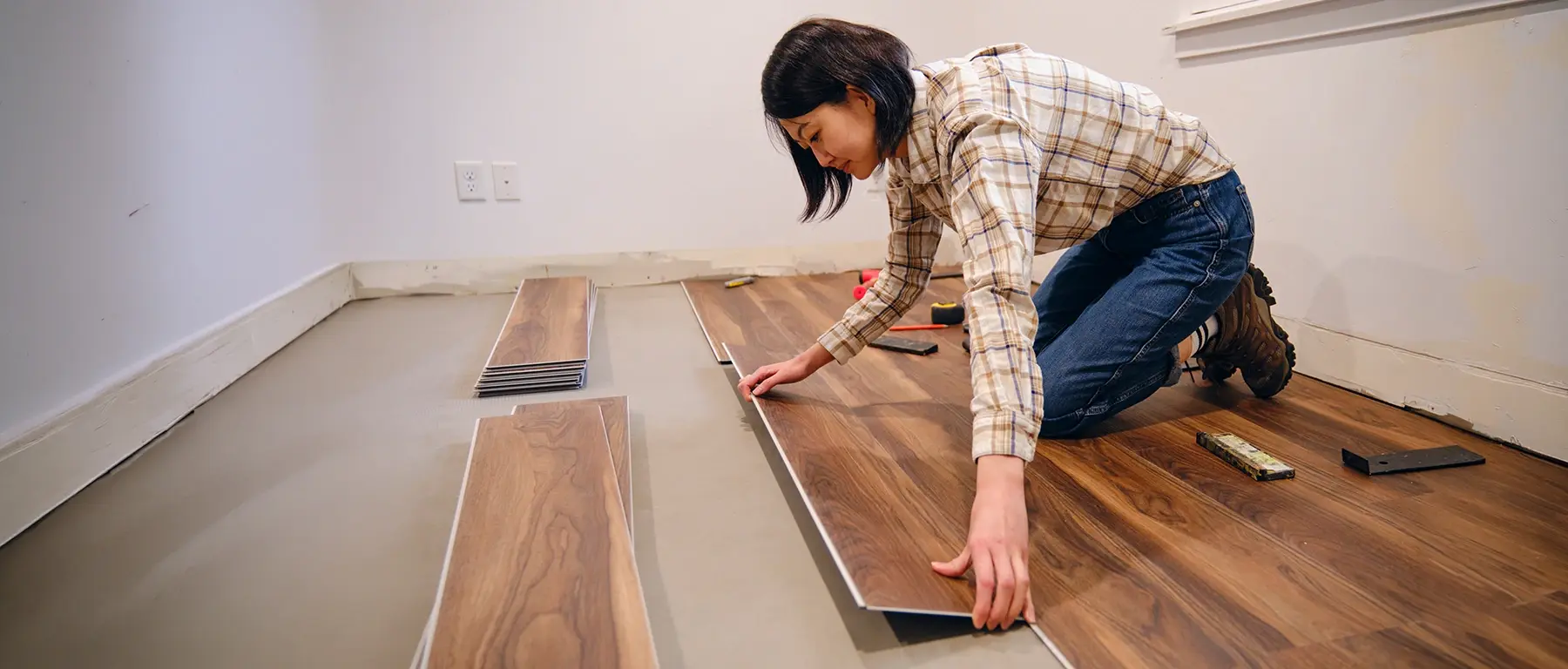 A woman in a plaid shirt kneels on a bare subfloor, measuring and laying wood-look plank flooring in a room under renovation.