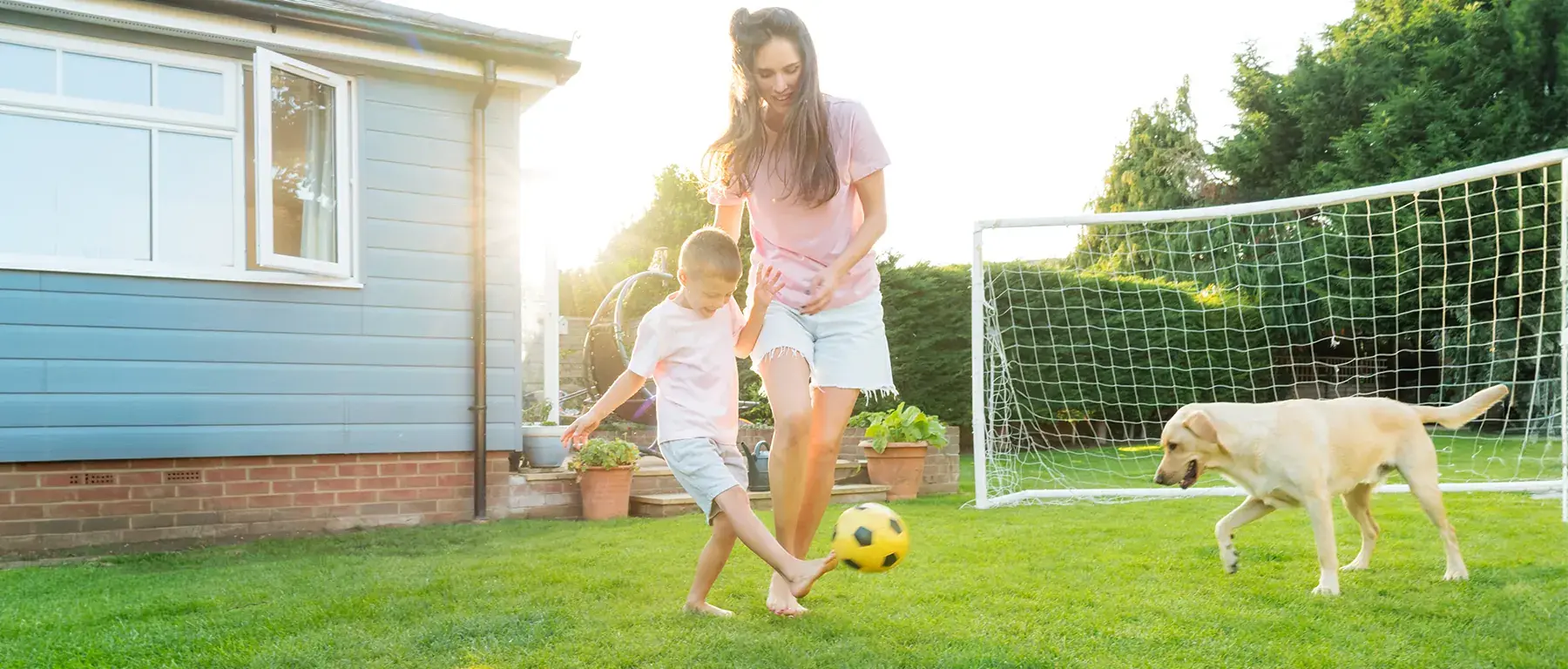 A woman and a child play soccer on a sunlit back lawn beside a pale blue clapboard house, with a goal net and a running yellow labrador