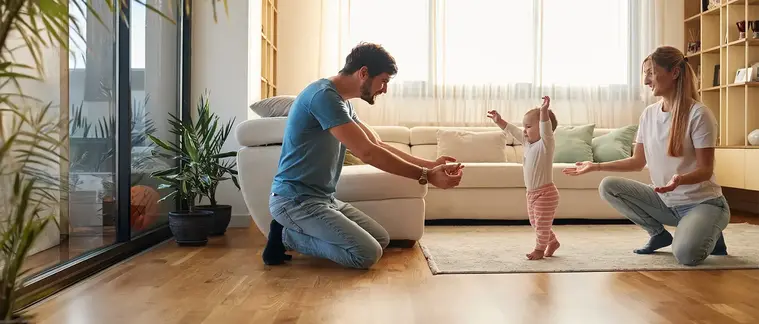 In a bright living room with wood floors, a man kneels with his arms open toward a toddler taking unsteady steps, while a woman crouches waiting on the other side