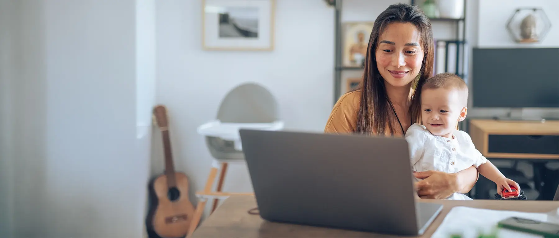 A woman sits at a wooden table working on a laptop while holding a baby on her lap, an acoustic guitar and a high chair behind her
