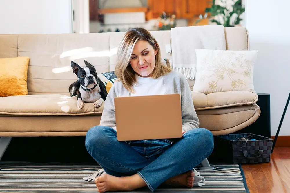 A woman sits cross-legged on the floor using a laptop while her small dog sits on the couch beside her.
