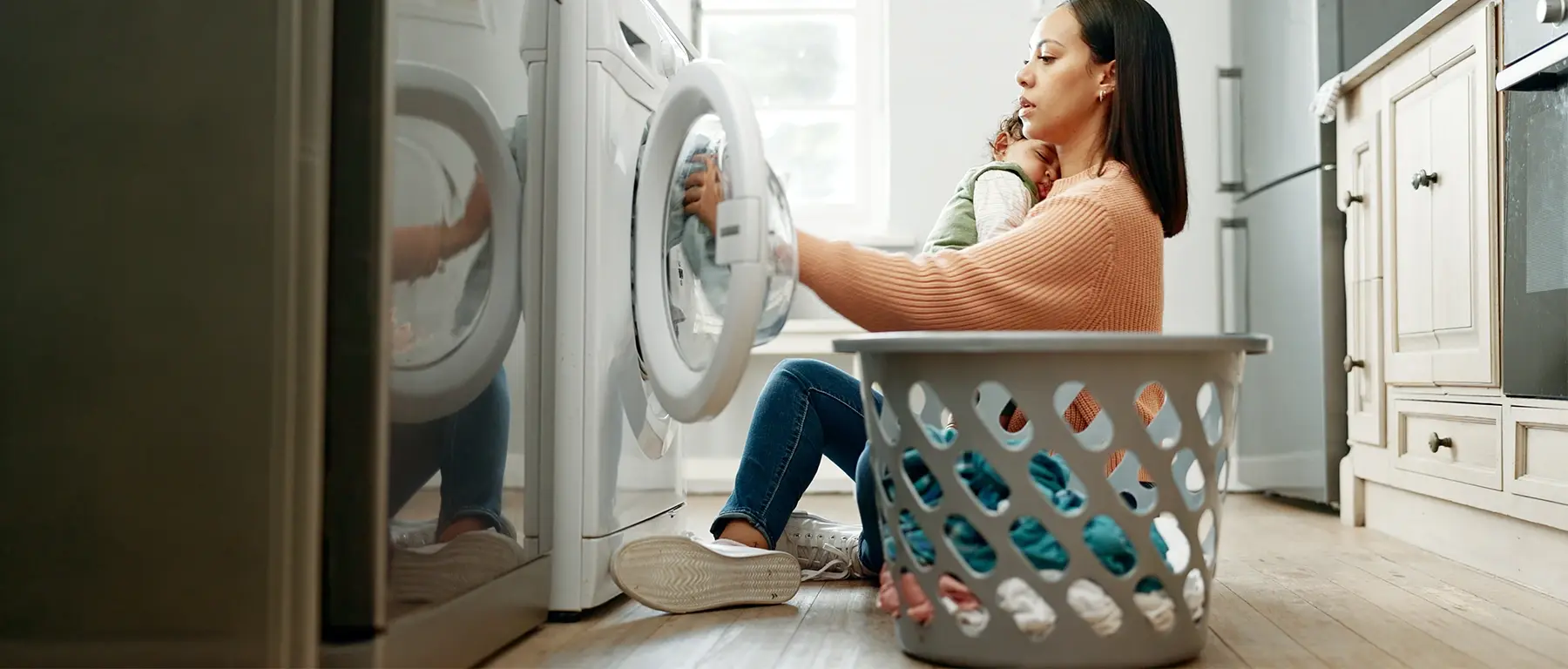 A woman kneels in front of an open front-loading washing machine in a kitchen, one hand on the door and a white laundry basket beside her