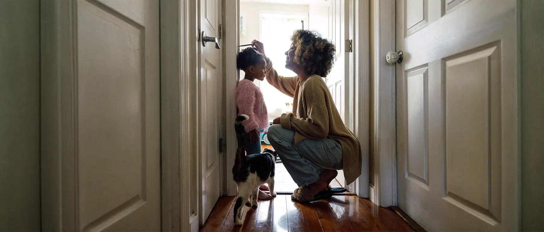 Seen down a hallway between two white doors, a woman crouches to help a child holding a toy in a doorway filled with bright daylight