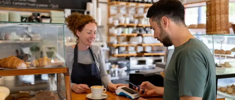 A customer taps to pay with a phone at a cafe counter while a barista assists.