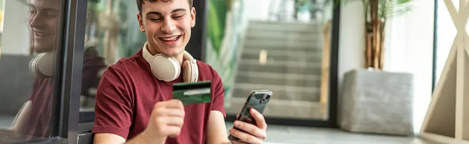 A young man with headphones around his neck smiles while holding a credit card and phone.