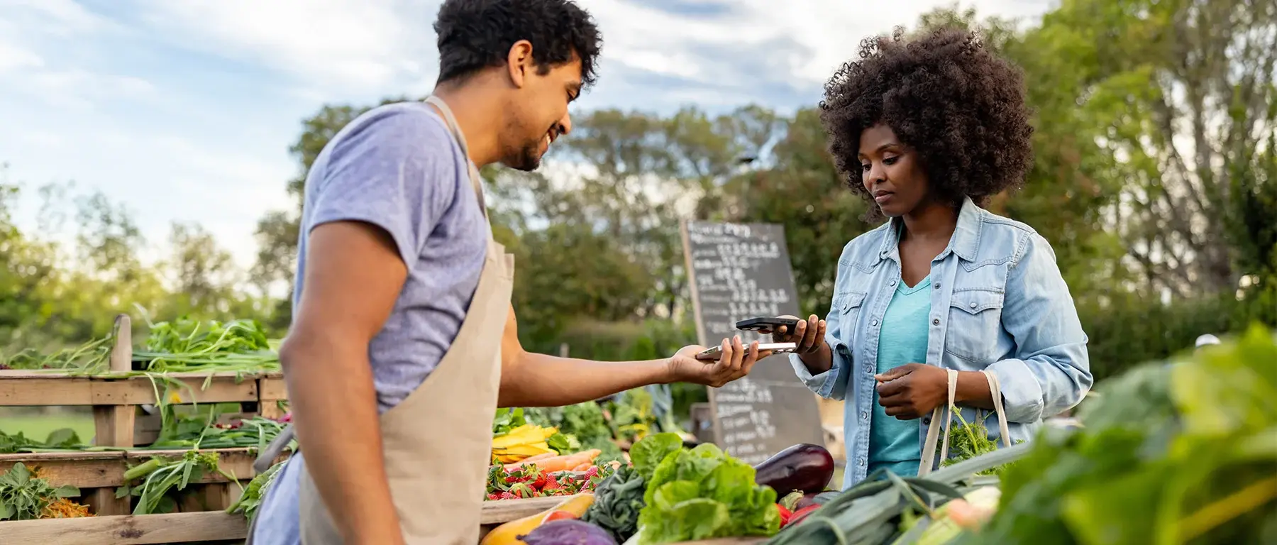 A farmers market vendor hands produce to a customer who taps to pay with her phone.