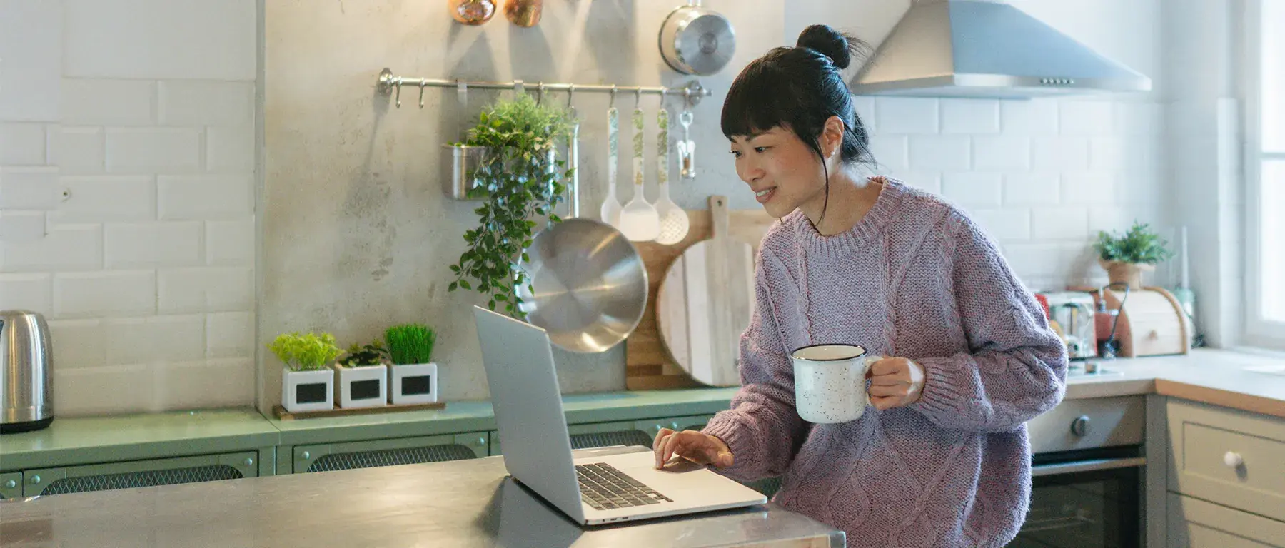 A woman stands at a bright kitchen counter with a mug, using a laptop open in front of her.