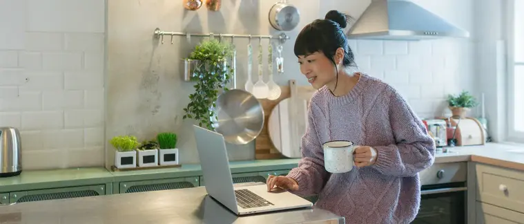 A woman stands at a bright kitchen counter with a mug, using a laptop open in front of her.