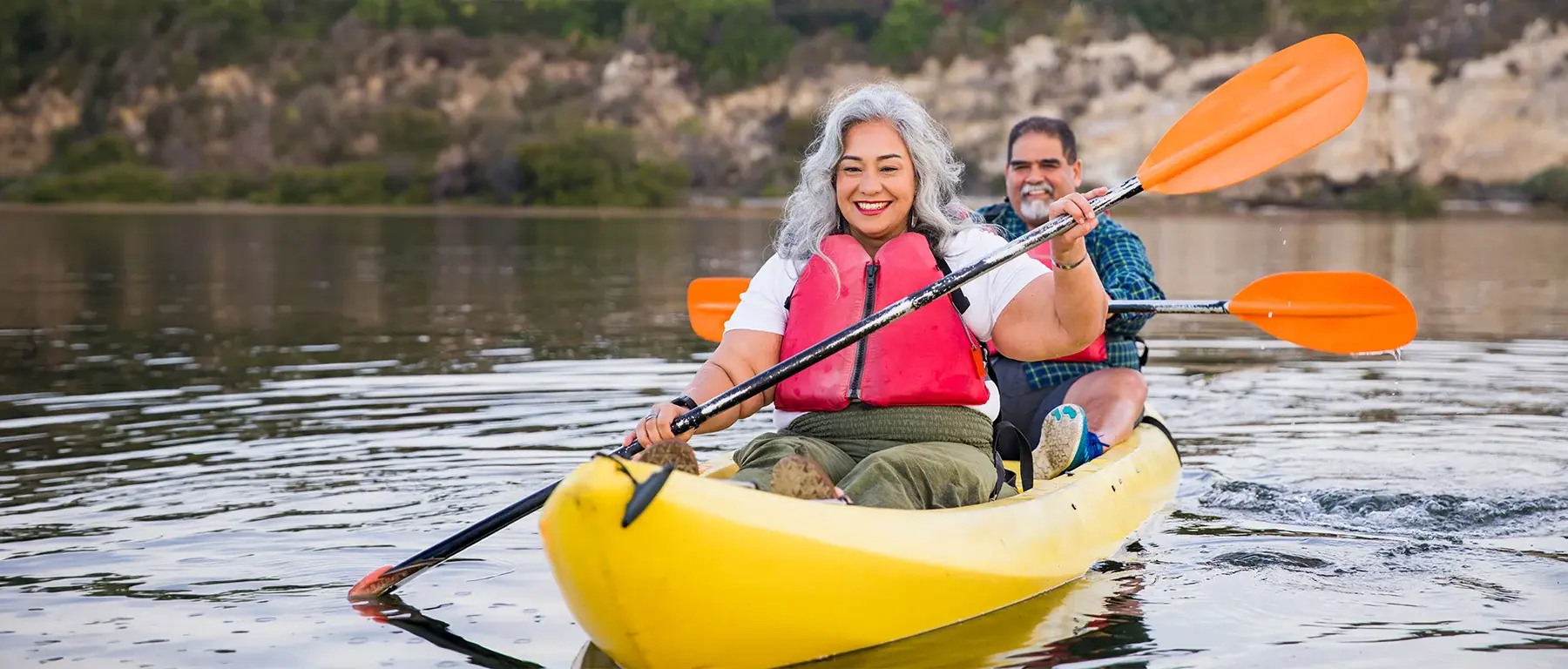 An older couple in life jackets smile while paddling a yellow kayak together on a calm lake.