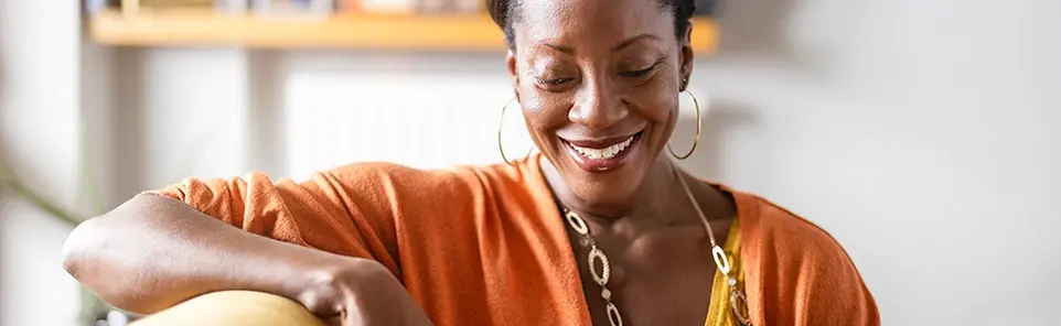 A smiling woman in an orange top relaxes at home, looking down at her phone.