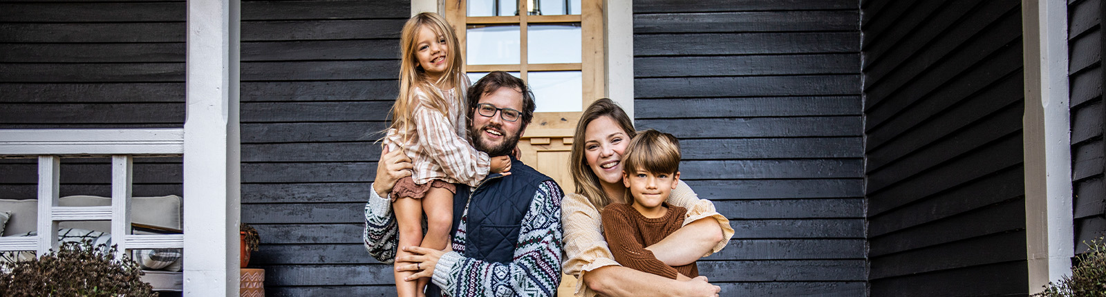 Two adults and two children sit together on the front steps of a dark clapboard house