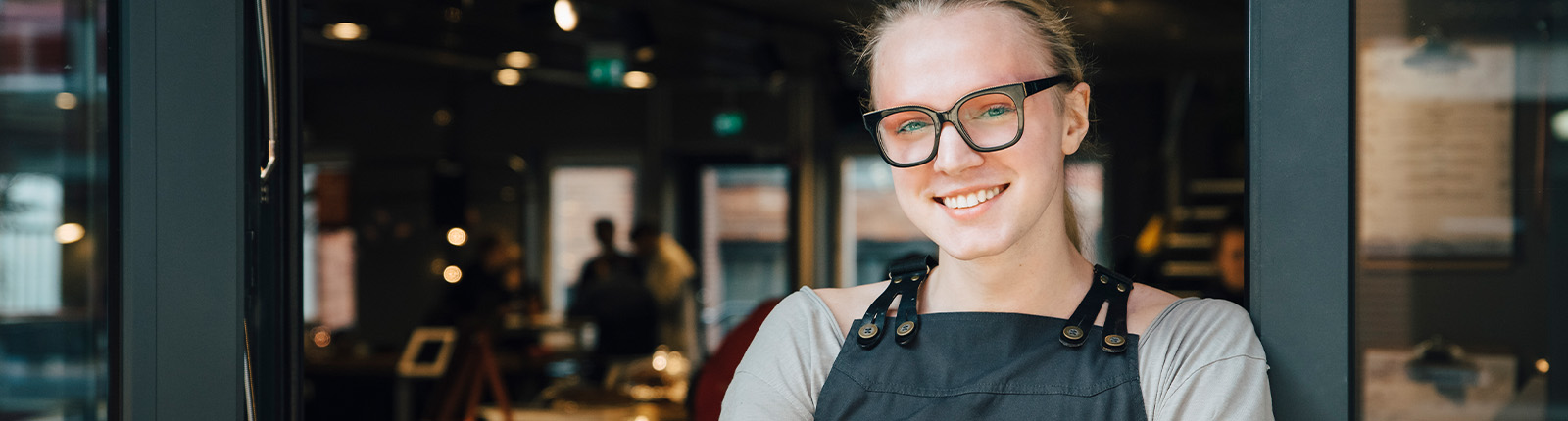 A person in an apron smiles in the doorway of a cafe, the lit interior behind them
