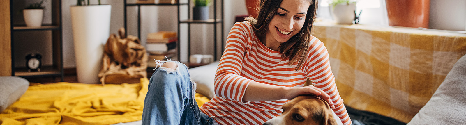 A person sits at home in a sunlit room, smiling and petting a dog beside them