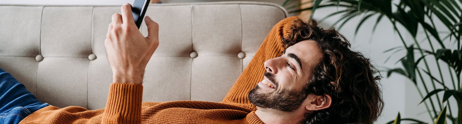A person lies back on a sofa smiling at their phone, a houseplant beside them
