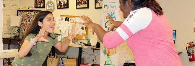 Two people laugh mid-gesture in a room hung with children's artwork and a hand-lettered poster