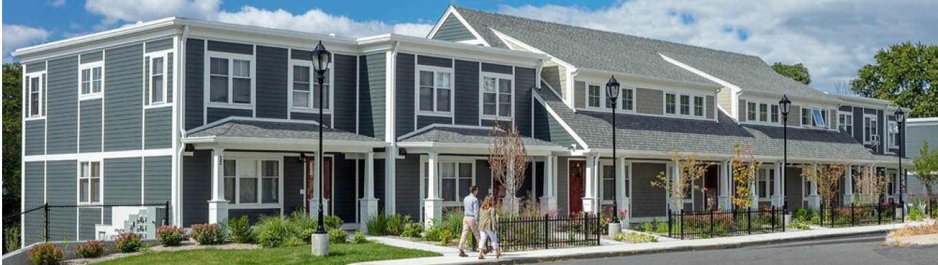 A row of new grey townhouses with porches and lamp posts, two people walking past