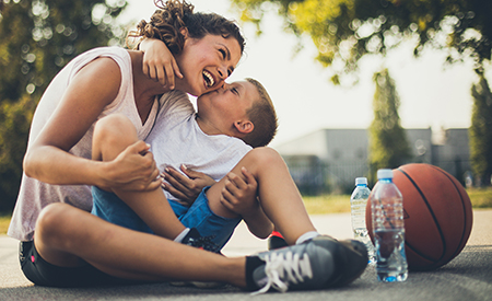 An adult and child hug and laugh on a basketball court, a ball and water bottles beside them
