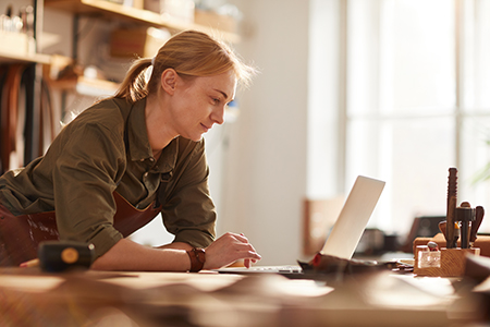 A person in a work apron leans over a laptop at a workbench in a sunlit workshop