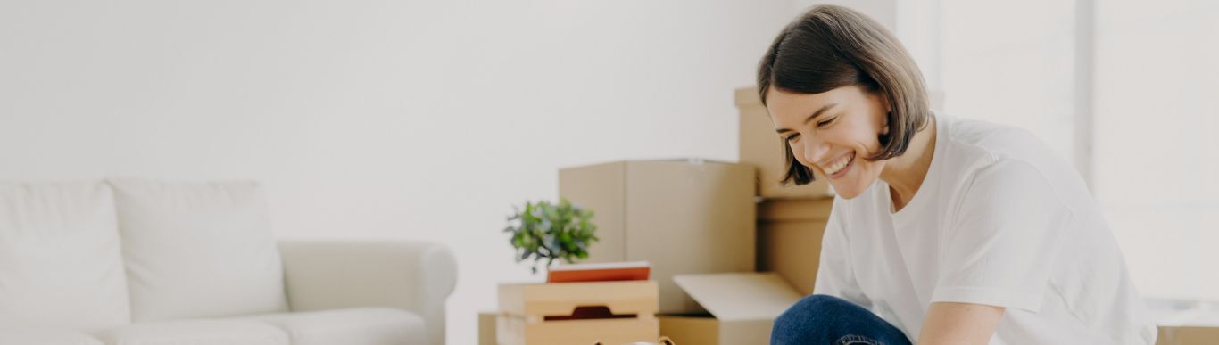 A person kneels smiling among moving boxes in a bright, nearly empty room