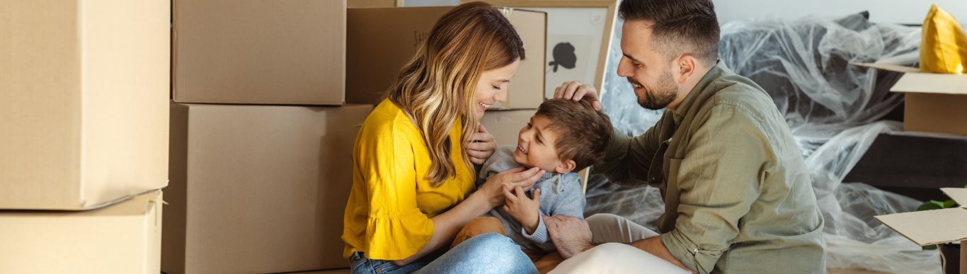 Two adults and a child sit laughing together on the floor among moving boxes