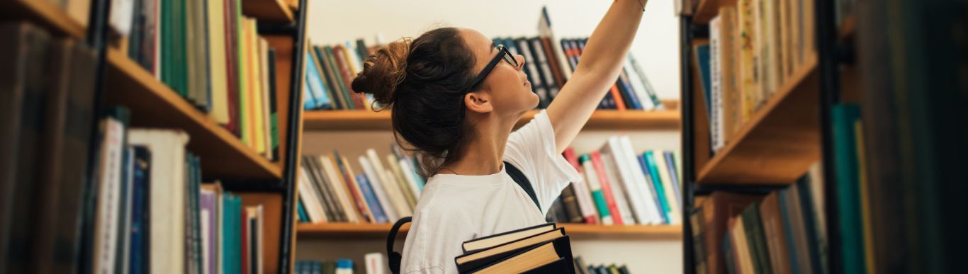 A student reaches for a book on a high library shelf, an armful of books already held