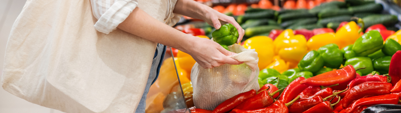 A shopper with a tote bag puts a green pepper into a mesh bag at a market produce display