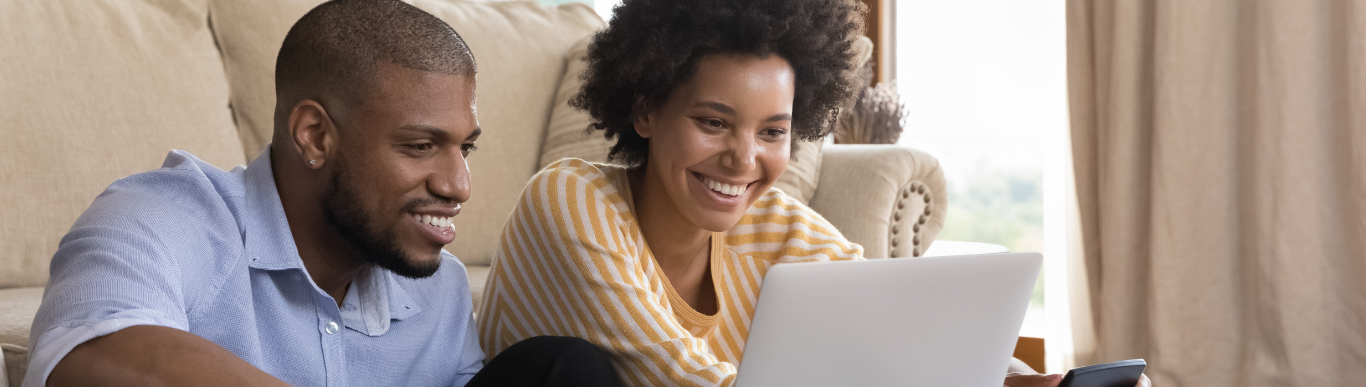 Two people sit on a sofa smiling at a laptop, one holding a phone