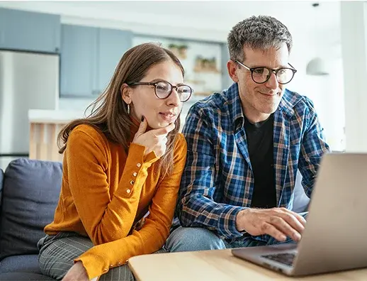 A woman and a man sit together on a sofa looking at a laptop screen in a bright kitchen.