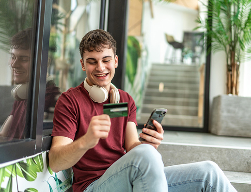 Young man in maroon t-shirt with headphones around neck smiling while holding smartphone and wallet in modern indoor setting with plants and stairs