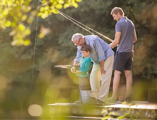 Three generations of a family fish together from a wooden dock on a sunlit lake.