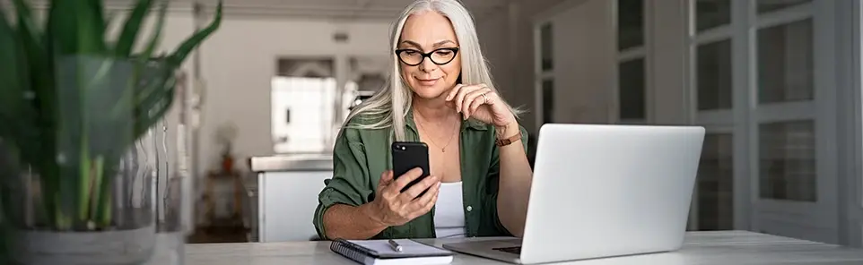 A woman with grey hair and glasses sits at a desk holding a phone, a laptop beside her.